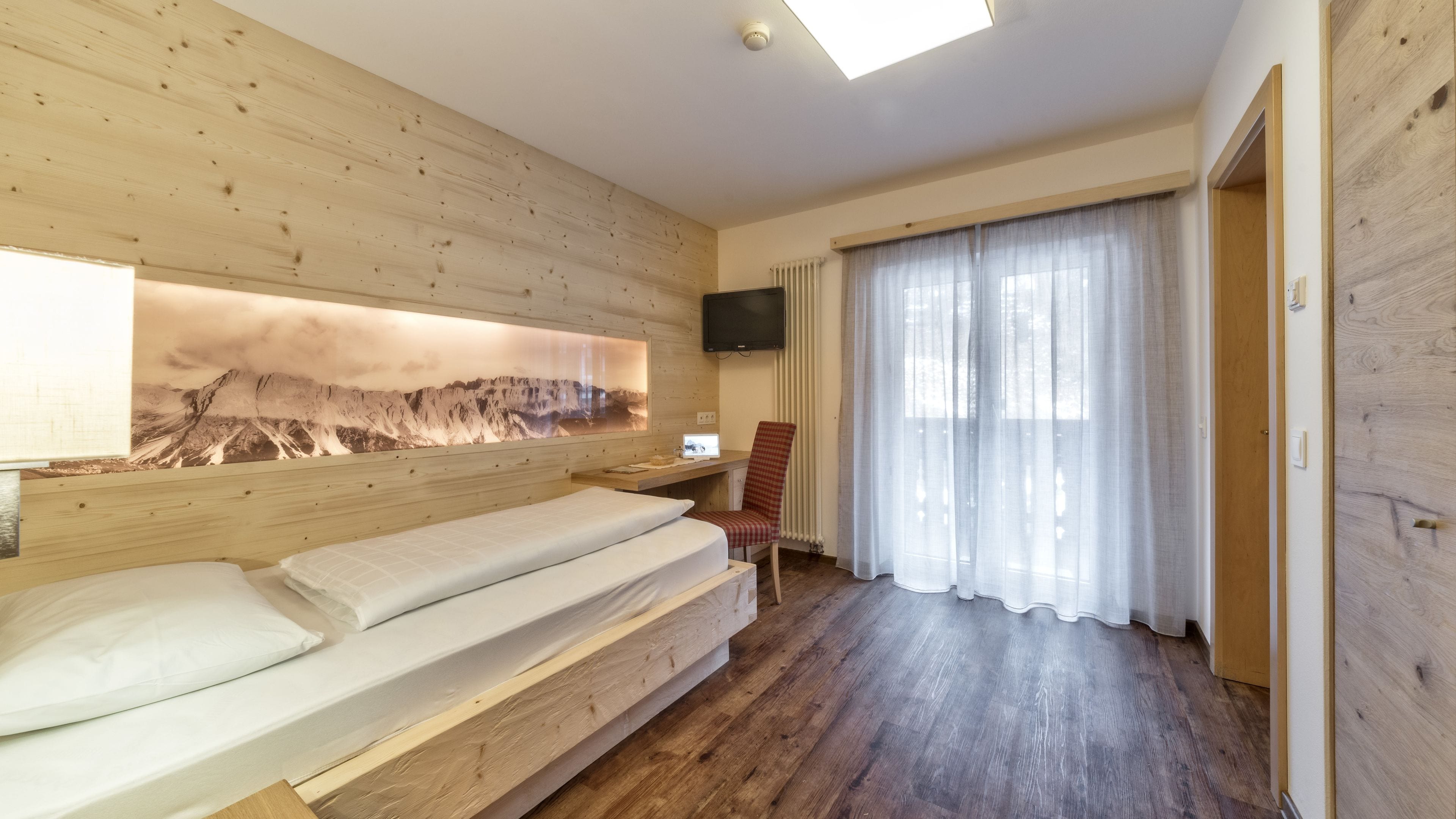 A room with a wooden bed, a desk with a chair, a mounted TV, and a large window with sheer curtains.
