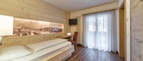 A room with a wooden bed, a desk with a chair, a mounted TV, and a large window with sheer curtains.