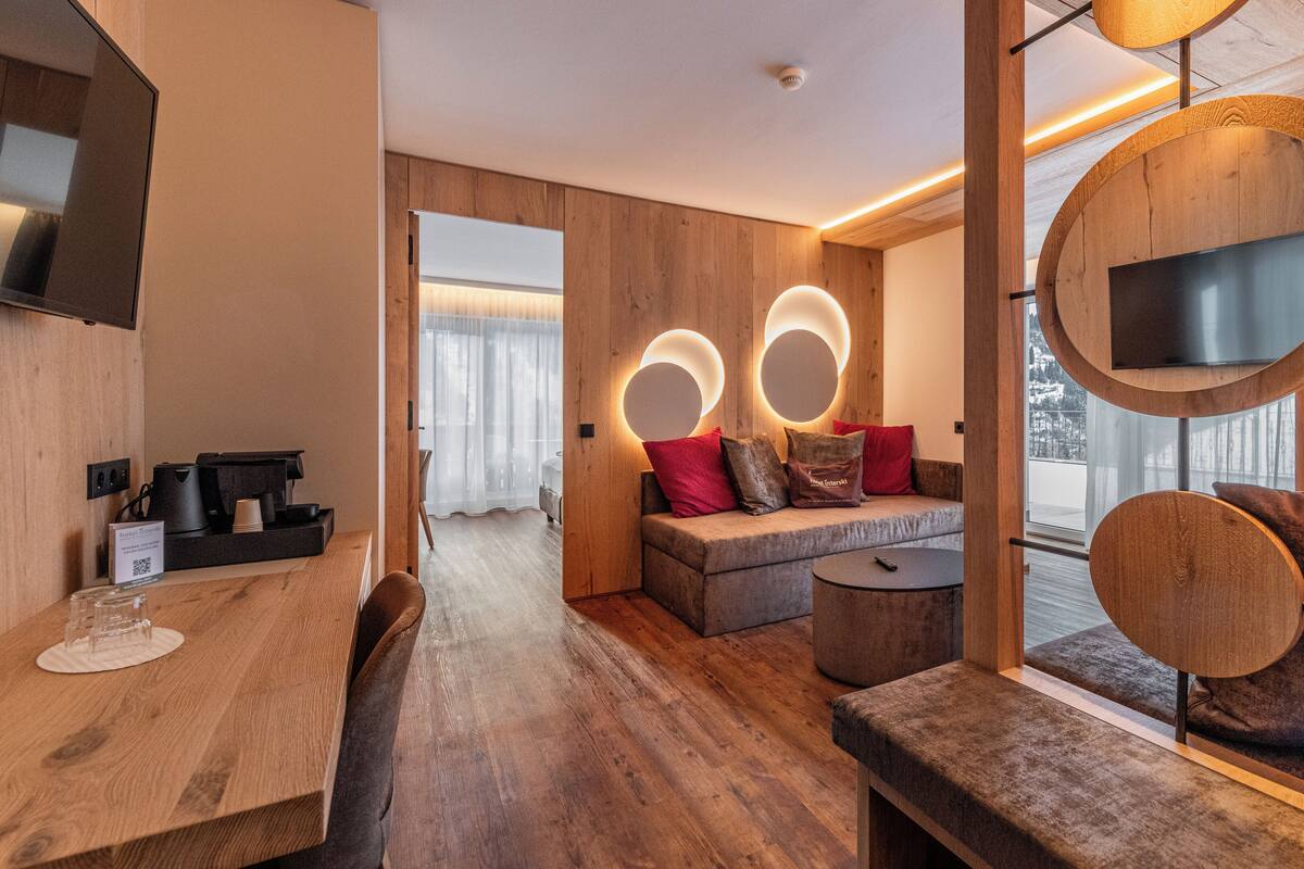 Una habitación de hotel moderna con suelo de madera, un sofá con cojines rojos, una mesa de centro redonda y un televisor montado en un panel de madera.