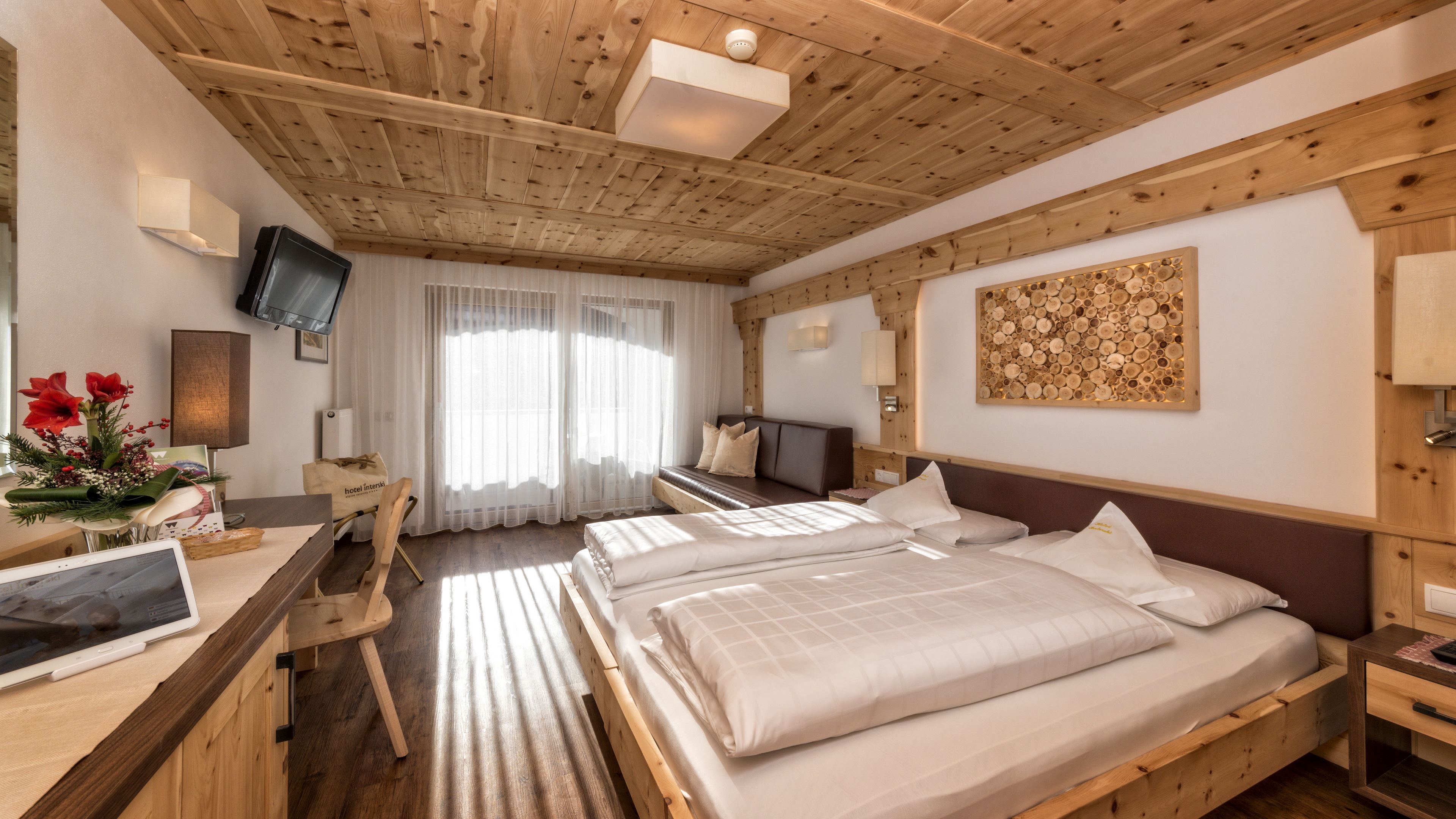 A hotel room with a wooden ceiling, a bed with white linens, a desk with a computer, and a TV on the wall.