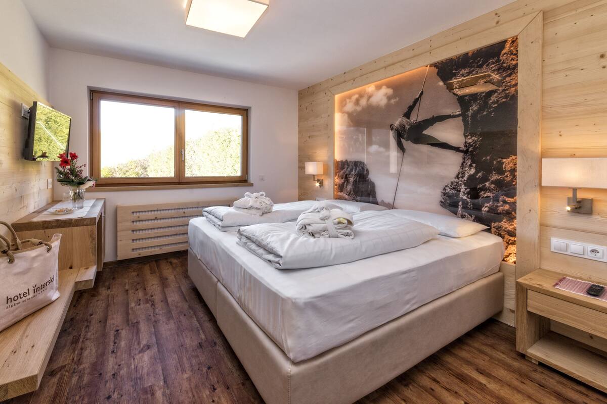 Un dormitorio con piso de madera, una cama con ropa de cama blanca, una ventana con vista, una lámpara de pared y un cabecero de madera con un cuadro.