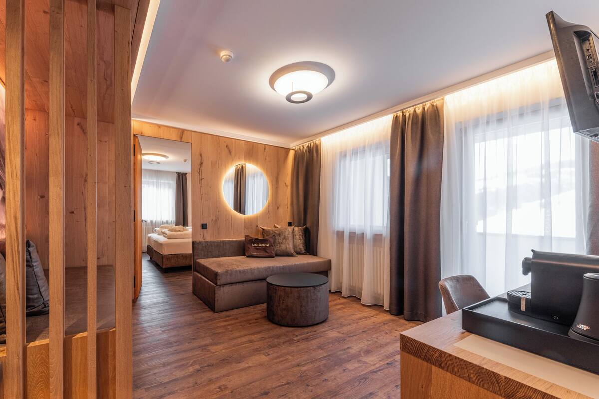 Una habitación de hotel moderna con piso de madera, un espejo redondo, un sofá, una mesa de centro y un televisor sobre un mueble de madera.