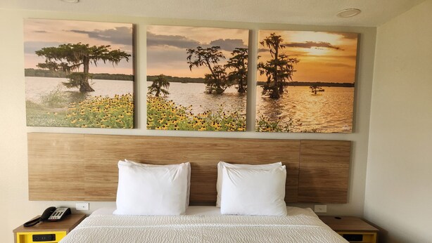 Camera d'albergo con un letto, due cuscini bianchi, una testiera in legno, un comodino con un telefono e una parete con tre dipinti raffiguranti un tramonto sull'acqua.