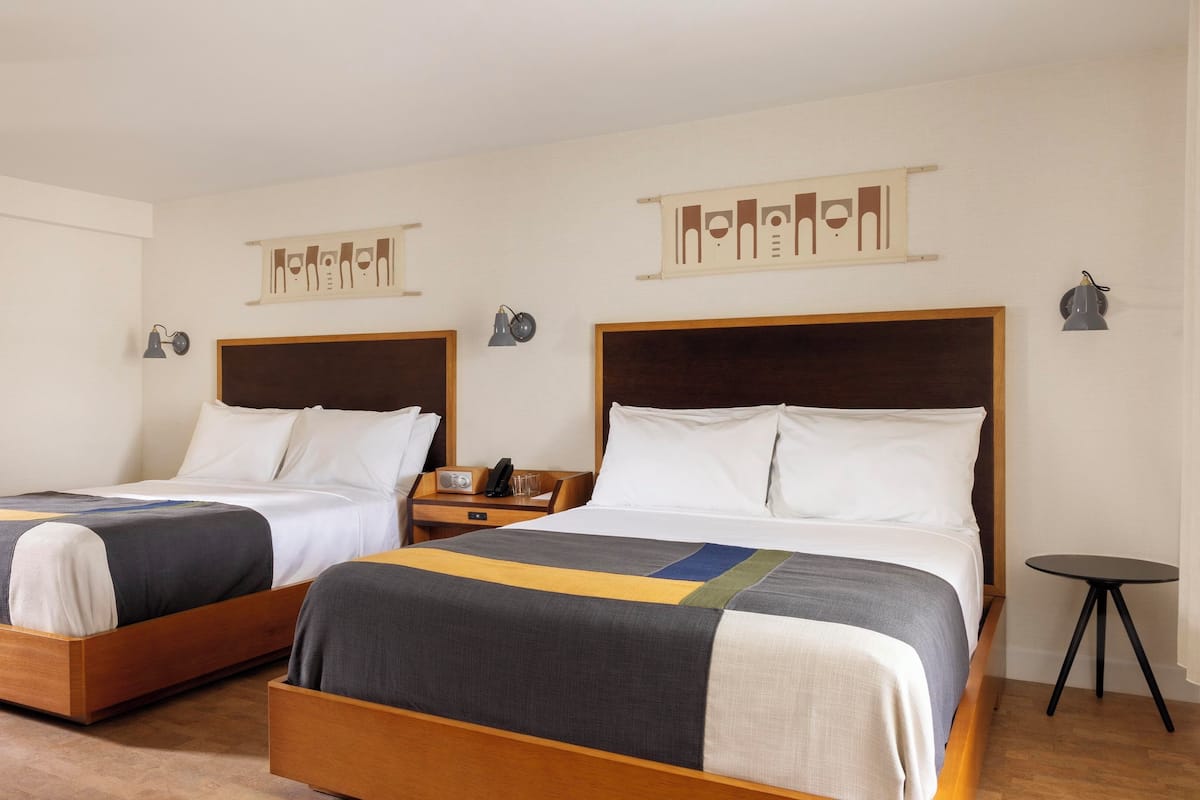 A hotel room with two beds, each with wooden frames and white bedding. There are bedside tables with lamps and a small table with a radio and glasses.