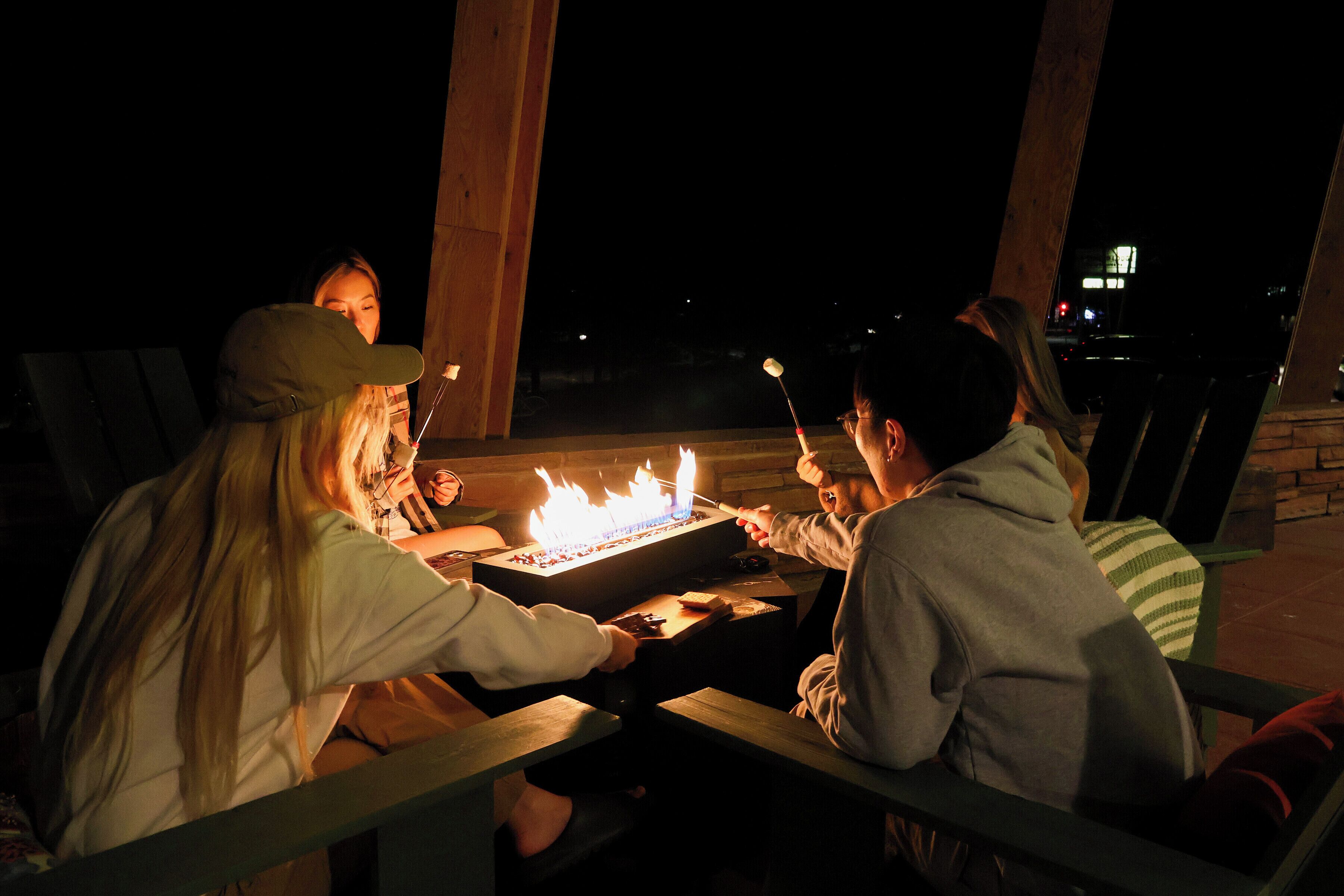 Un groupe de personnes est assis autour d’une table en bois, avec un foyer au centre, à l’extérieur, pendant la nuit.
