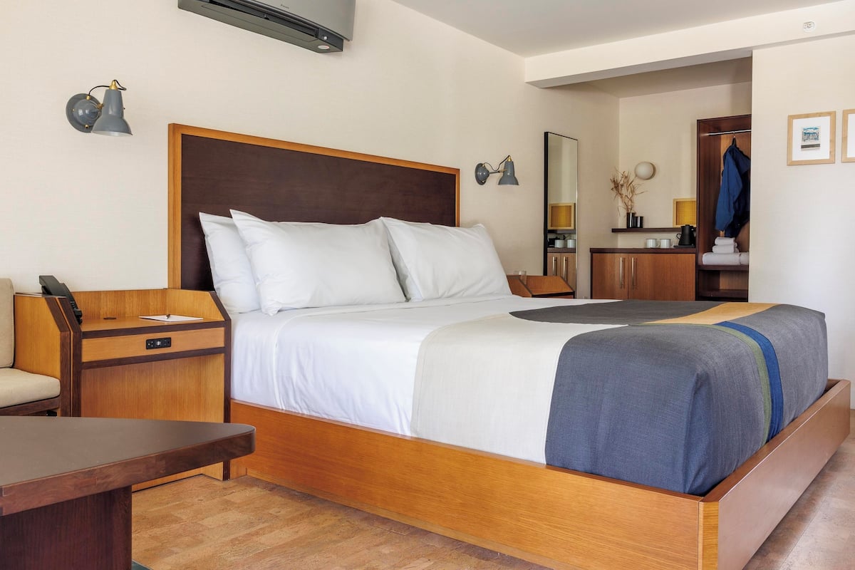 A hotel room with a wooden bed, a bedside table, a chair, a desk, and a small kitchenette area.