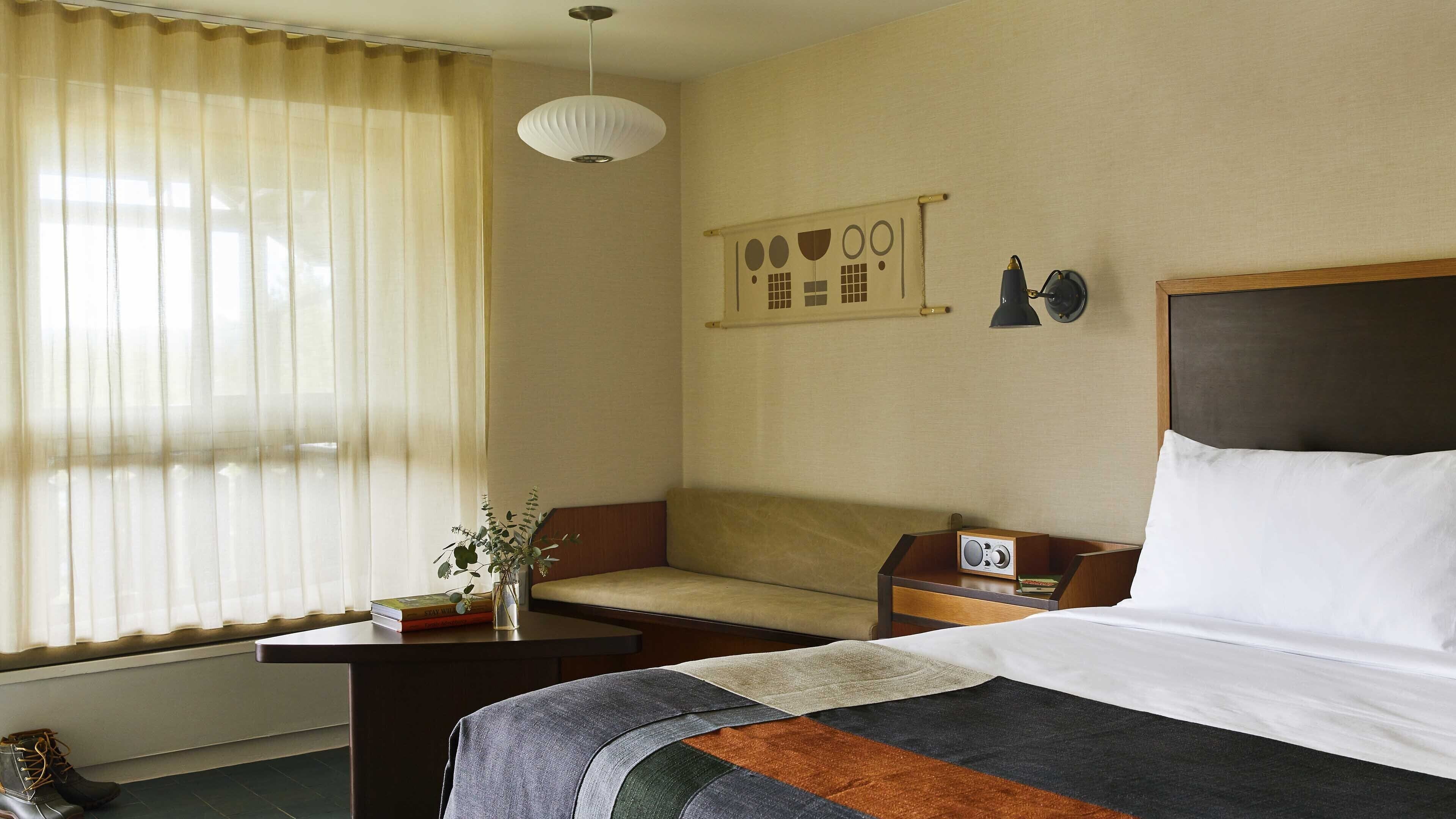Ein Hotelzimmer mit einem Bett, einem Sofa, einem kleinen Tisch mit Lampe, einer Wanduhr und einem Fenster mit Vorhängen.