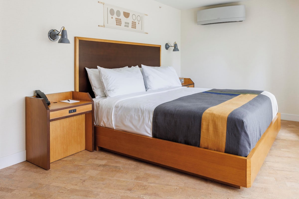 A modern hotel room with a wooden bed, a nightstand with a phone, and a wall-mounted air conditioner.