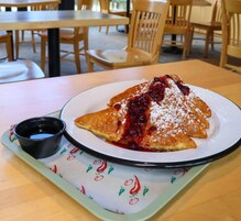 Un plato de tortitas con jarabe y azúcar glas, acompañado de una taza pequeña de jarabe, sobre una mesa con posavasos.