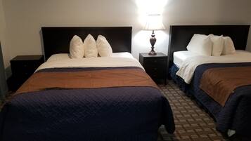 A hotel room with two beds, each with white bedding and a brown blanket. There is a nightstand with a lamp between the beds.