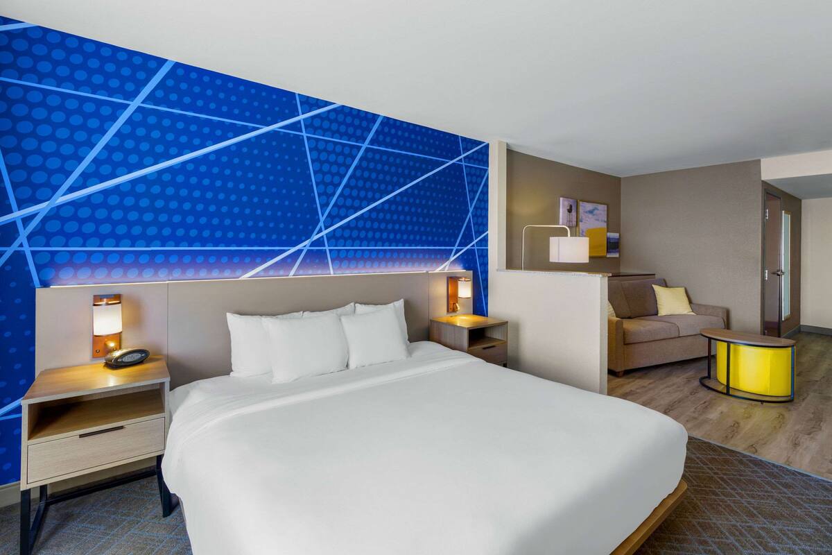 A modern hotel room with a large bed, a nightstand with a lamp, a small table with a lamp, a sofa, and a wall with a geometric pattern.