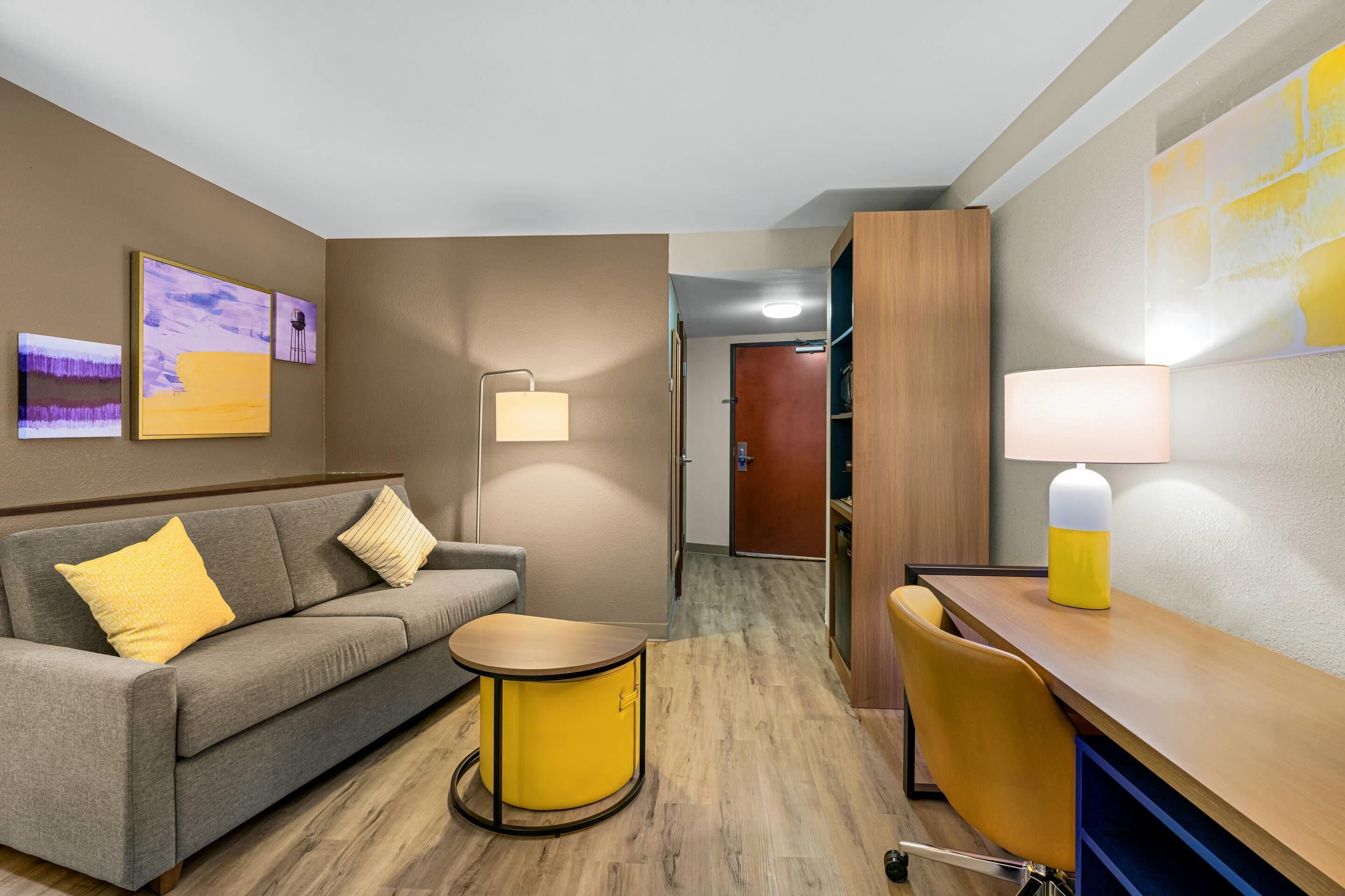 A hotel room with a sofa, a small round table, a desk with a chair, a lamp, and artwork on the walls.