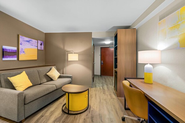 Una habitación de hotel con sofá, una mesita redonda, escritorio con silla, lámpara y cuadros en las paredes.