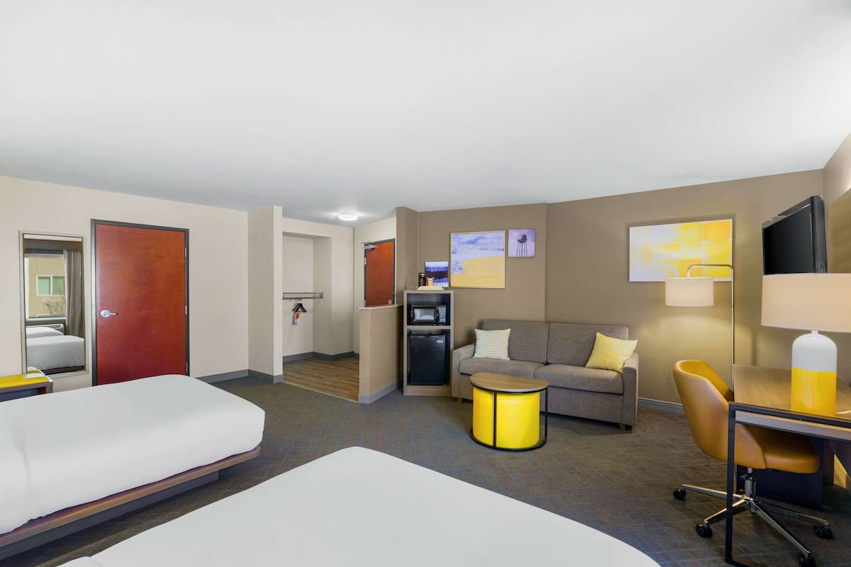 A hotel room with two beds, a sofa, a small table, a chair, a desk, a lamp, a TV, a microwave, and a refrigerator.