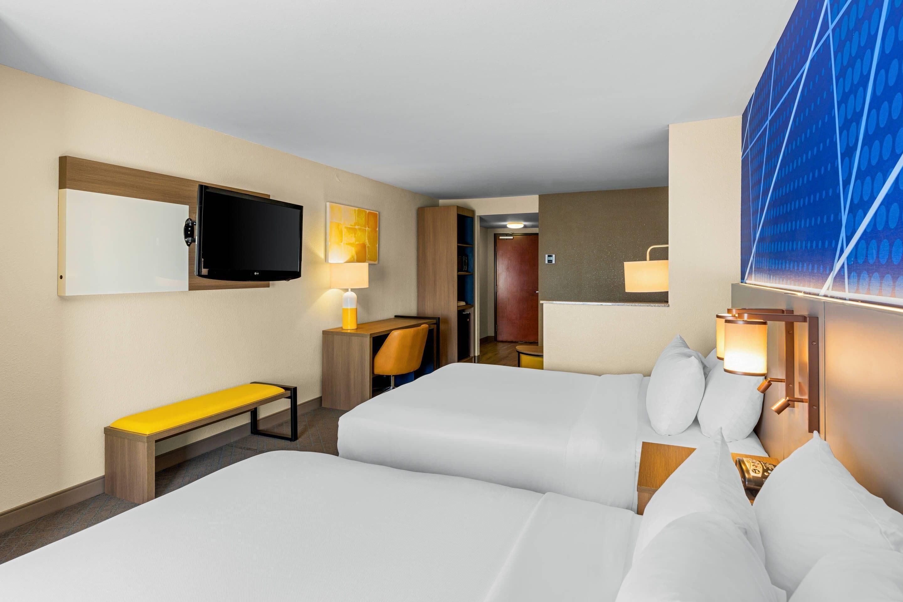 A hotel room with two beds, a desk with a chair, a television, and a large window with a view.