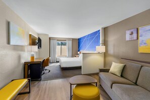 A hotel room with a bed, a sofa, a desk with a chair, a small table, and a TV mounted on the wall.