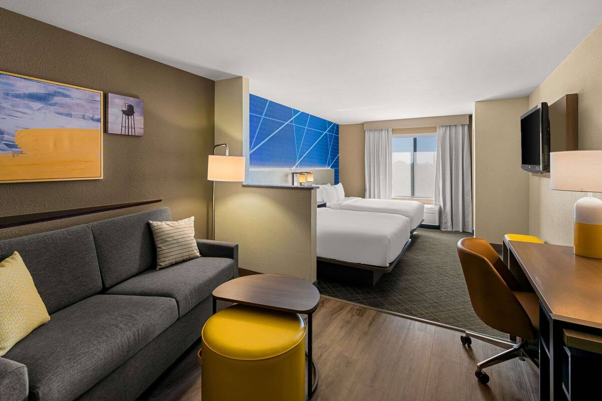 A hotel room with a sofa, a small table, a yellow ottoman, a bed, a desk, and a TV.