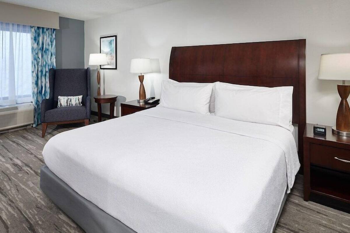 A hotel room with a large bed, a chair, two bedside tables with lamps, a window with curtains, and a wall-mounted air conditioner.