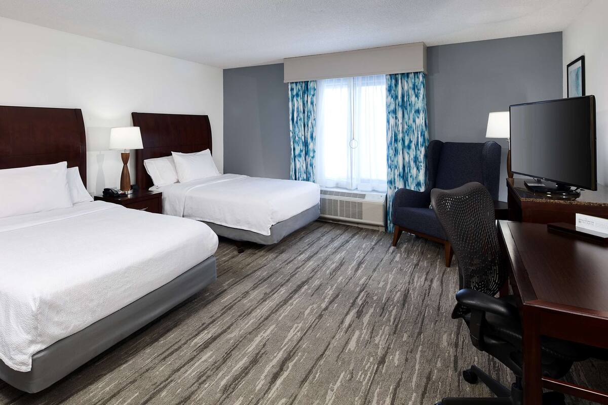 Ett hotellrum med två sängar, ett skrivbord med en stol, en tv och ett fönster med gardiner.