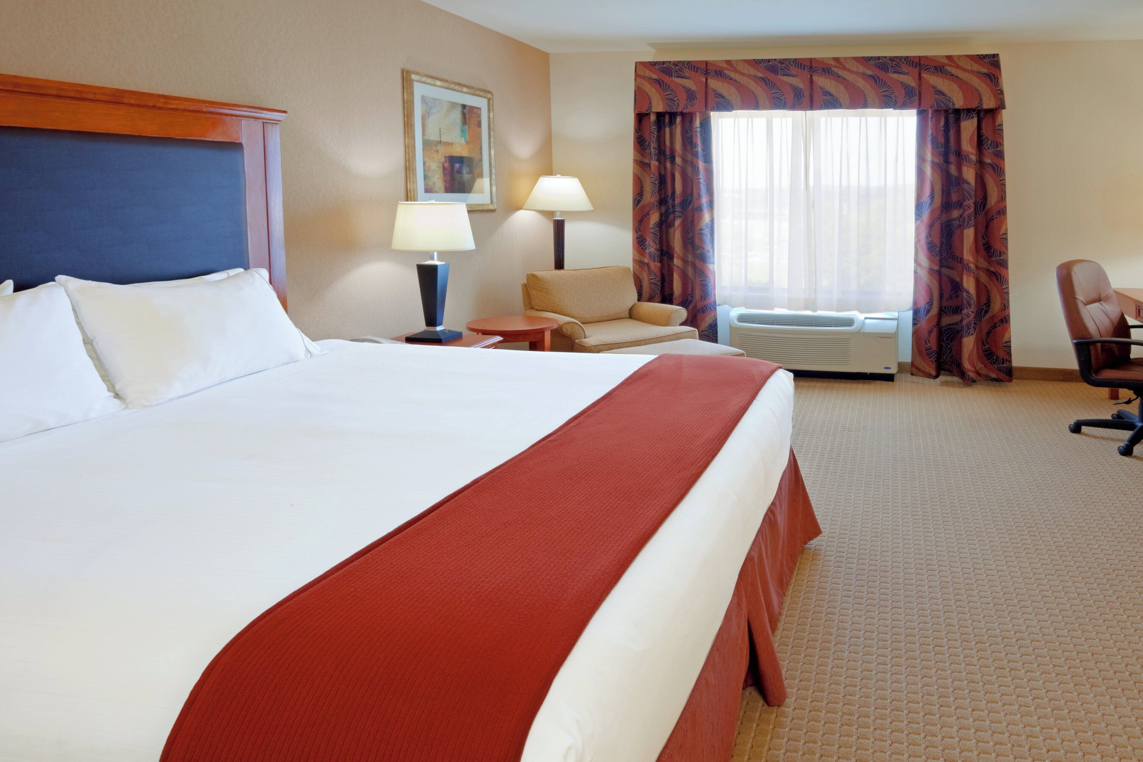 A hotel room with a large bed, a desk, a chair, a window with curtains, and an air conditioning unit.