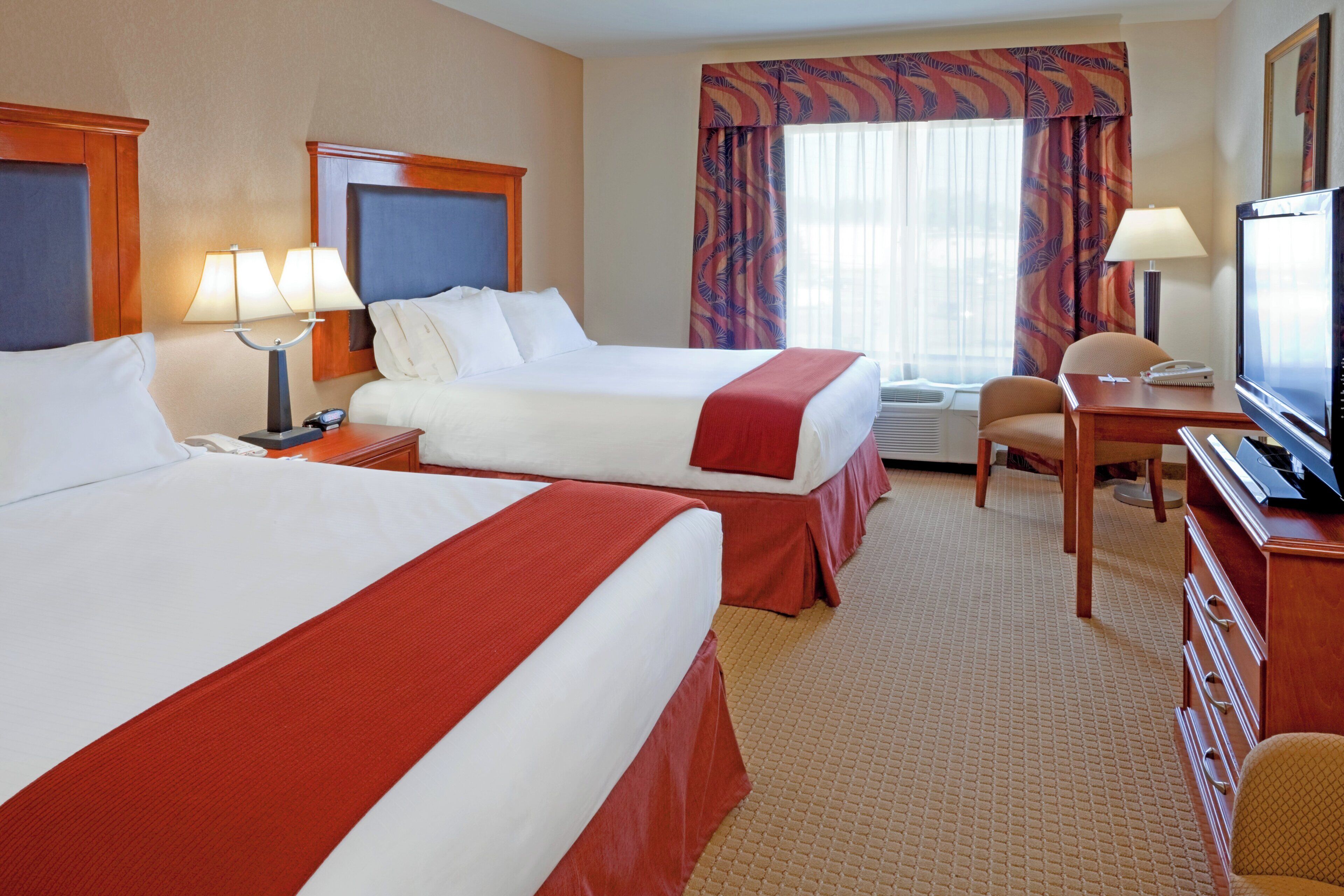 A hotel room with two beds, a desk with a television, a chair, and a window with curtains.