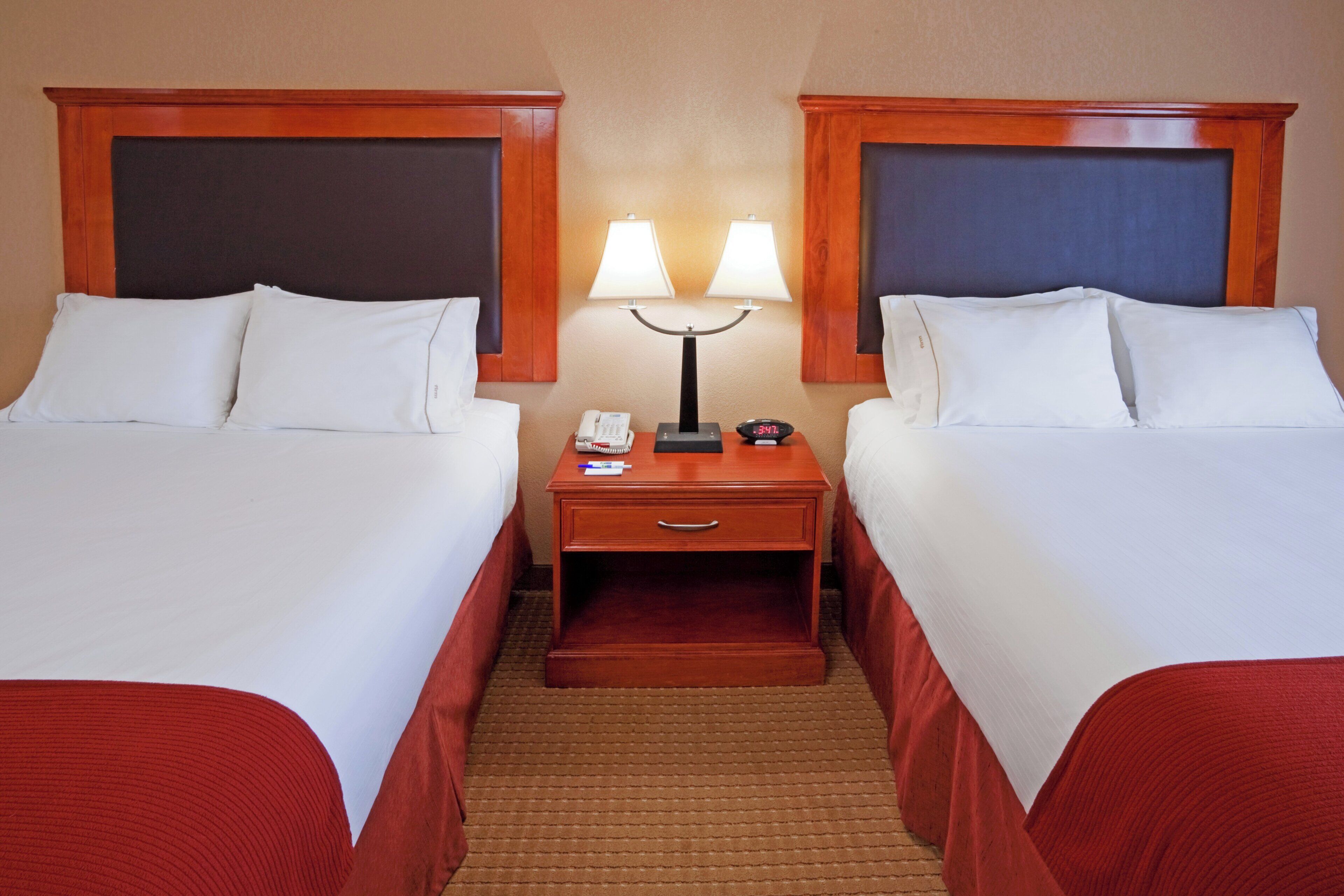 A hotel room with two single beds, a nightstand with a lamp, a phone, and a digital clock.