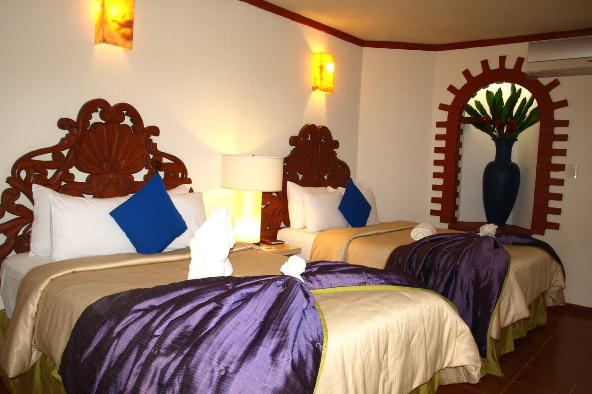 A hotel room with three beds, each with a wooden headboard, white and blue pillows, and a purple blanket. There is a large mirror with a decorative frame and a potted plant on the wall.