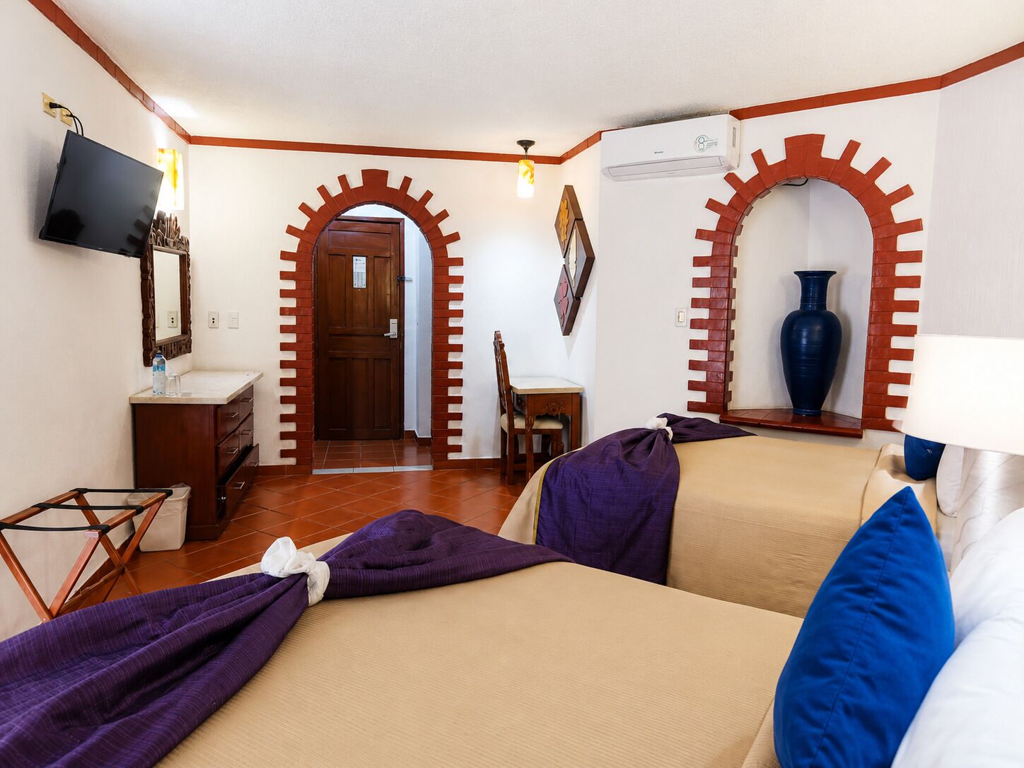 Habitación de hotel con cama, escritorio, silla, televisión, espejo, armario y un arco de entrada.