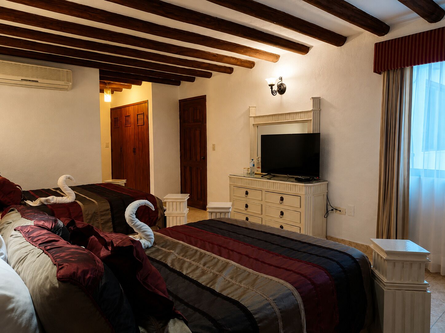 A hotel room with a bed, a television on a dresser, a window with curtains, and wooden beams on the ceiling.