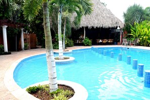A swimming pool with a thatched-roof structure in the background, surrounded by palm trees and potted plants. - Ecotel Quinta Regia (Valladolid)
