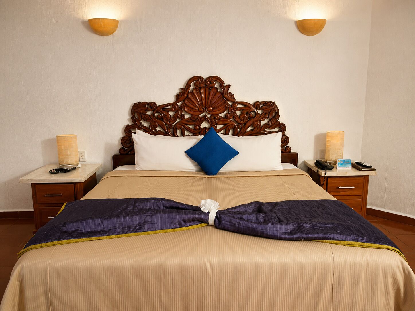 Une chambre d’hôtel avec un grand lit, une tête de lit en bois, deux tables de chevet avec des lampes et une petite table de nuit avec un téléphone et une carte.