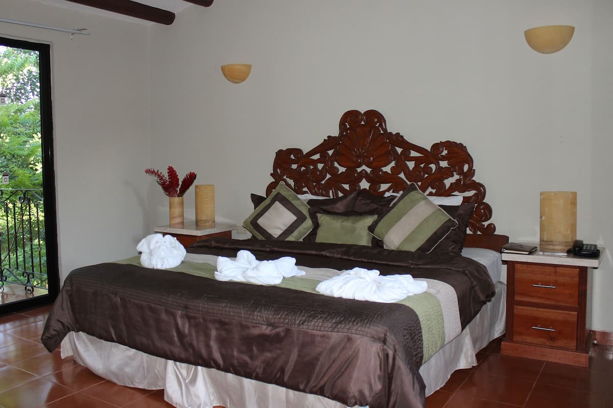 A neatly made bed with a brown headboard, a bedside table with a lamp, and a view of greenery outside.