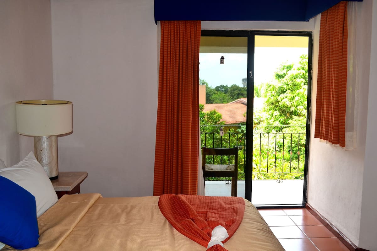 A hotel room with a bed, a nightstand with a lamp, and a view of a balcony with greenery.