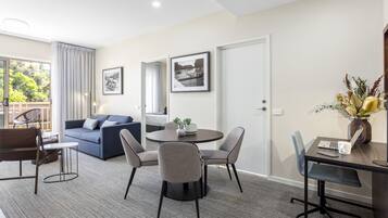 A modern living room with a sofa, dining table, and chairs, a balcony with a view, and a door with a framed picture.