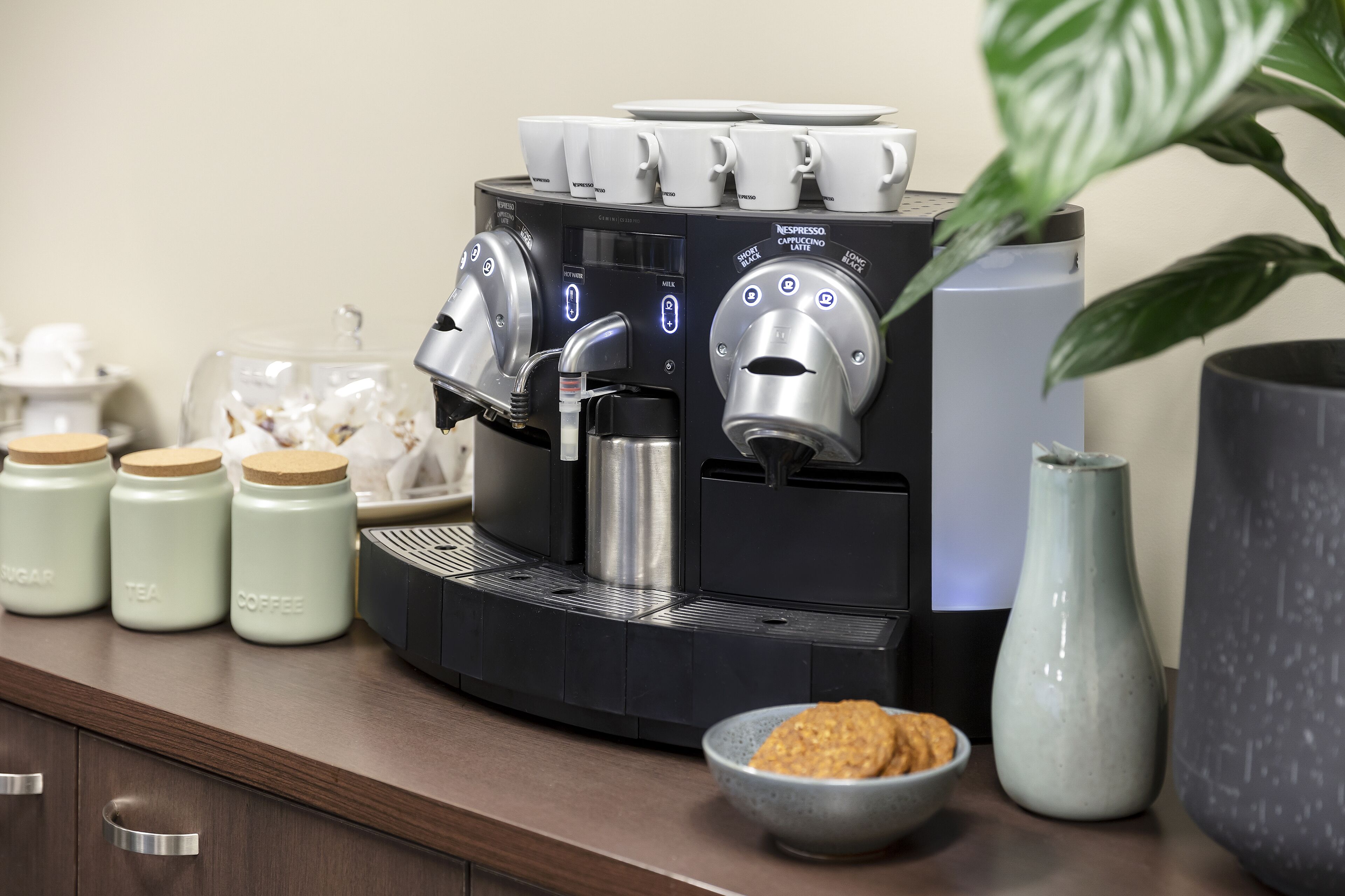 A coffee machine with multiple dispensers, a bowl of cookies, and jars labeled sugar, tea, and coffee on a counter.