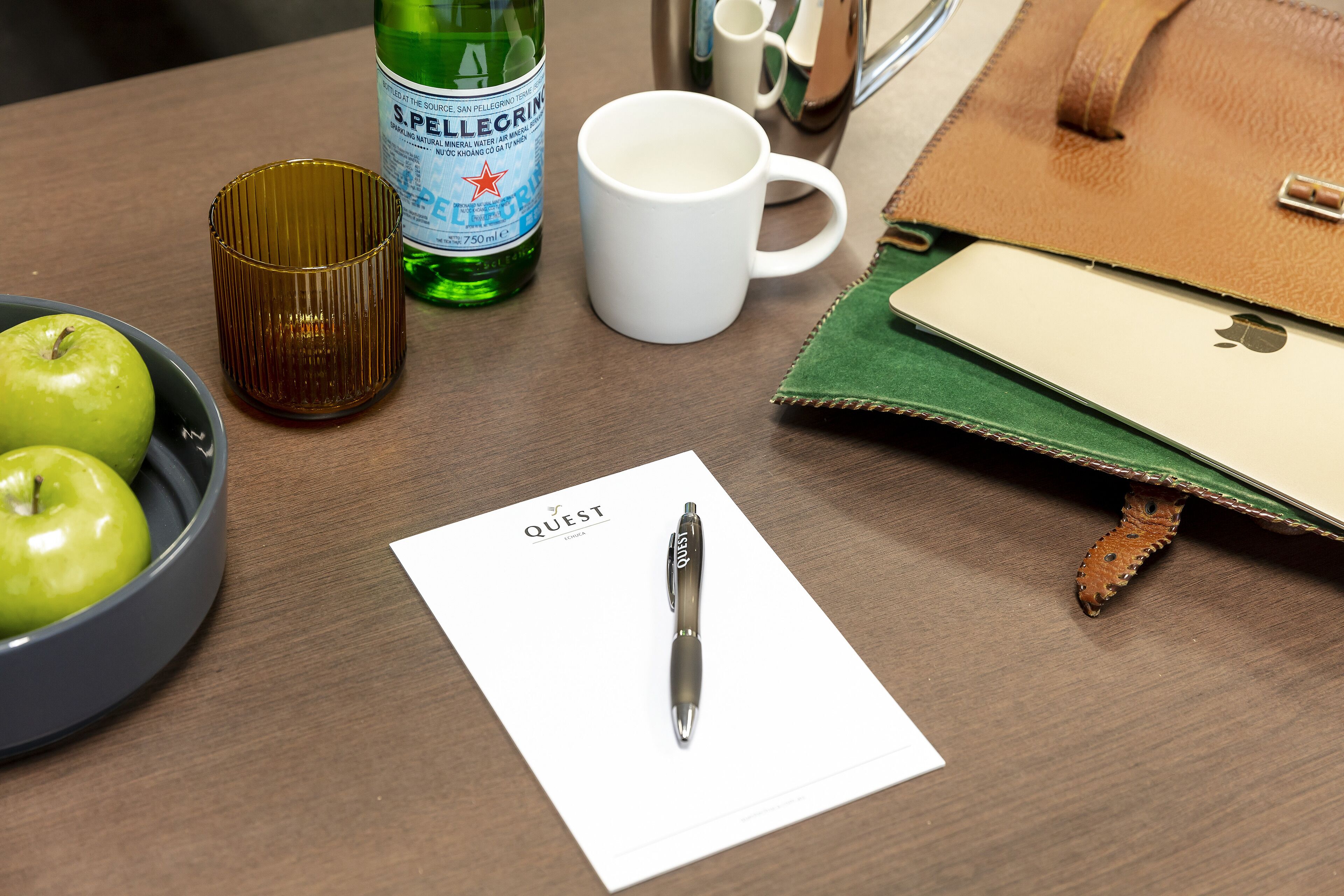 A desk with a bowl of green apples, a white mug, a green bottle, a pen, and a notebook.
