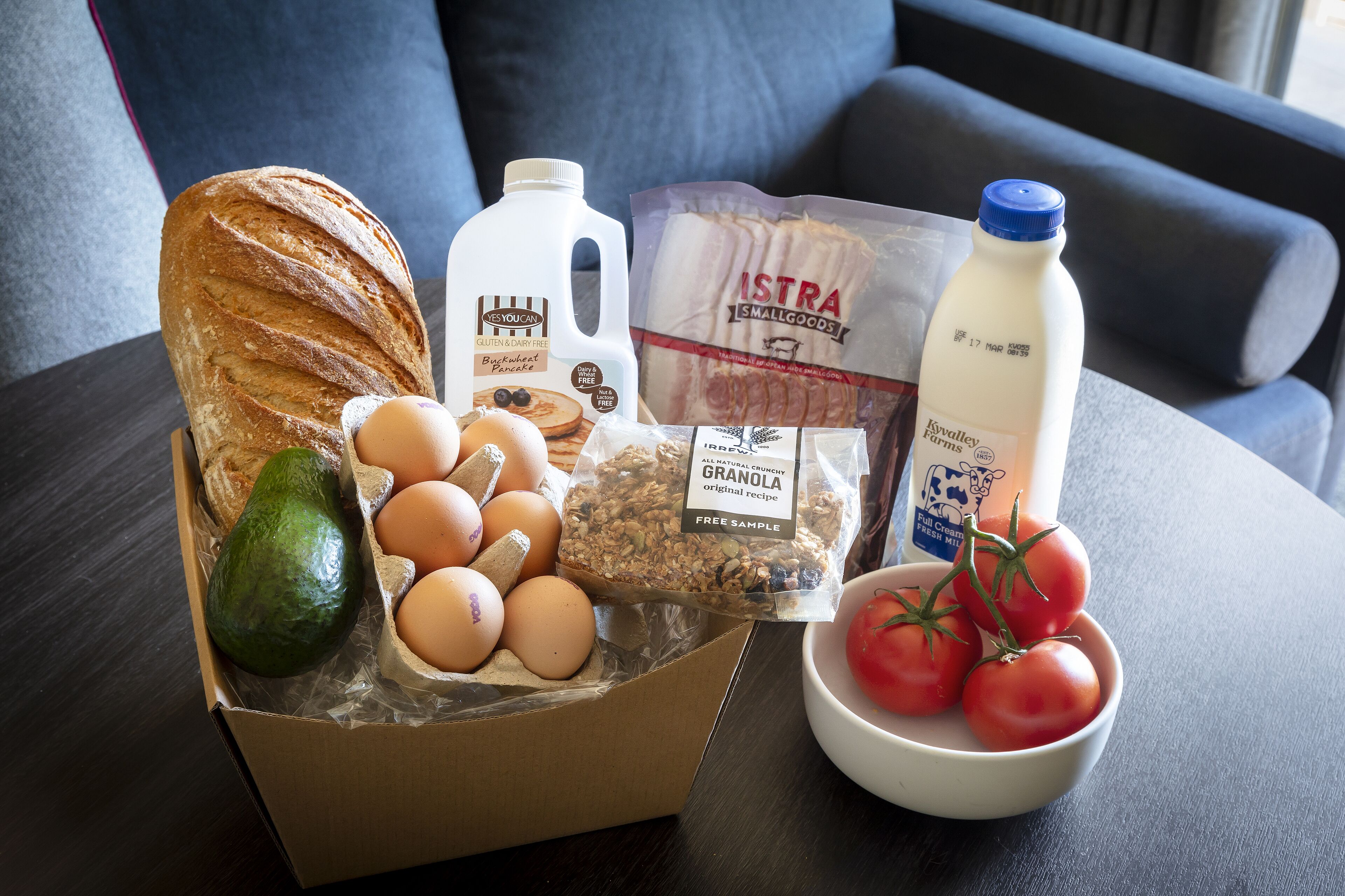 A box of groceries including bread, eggs, tomatoes, milk, and granola on a table.
