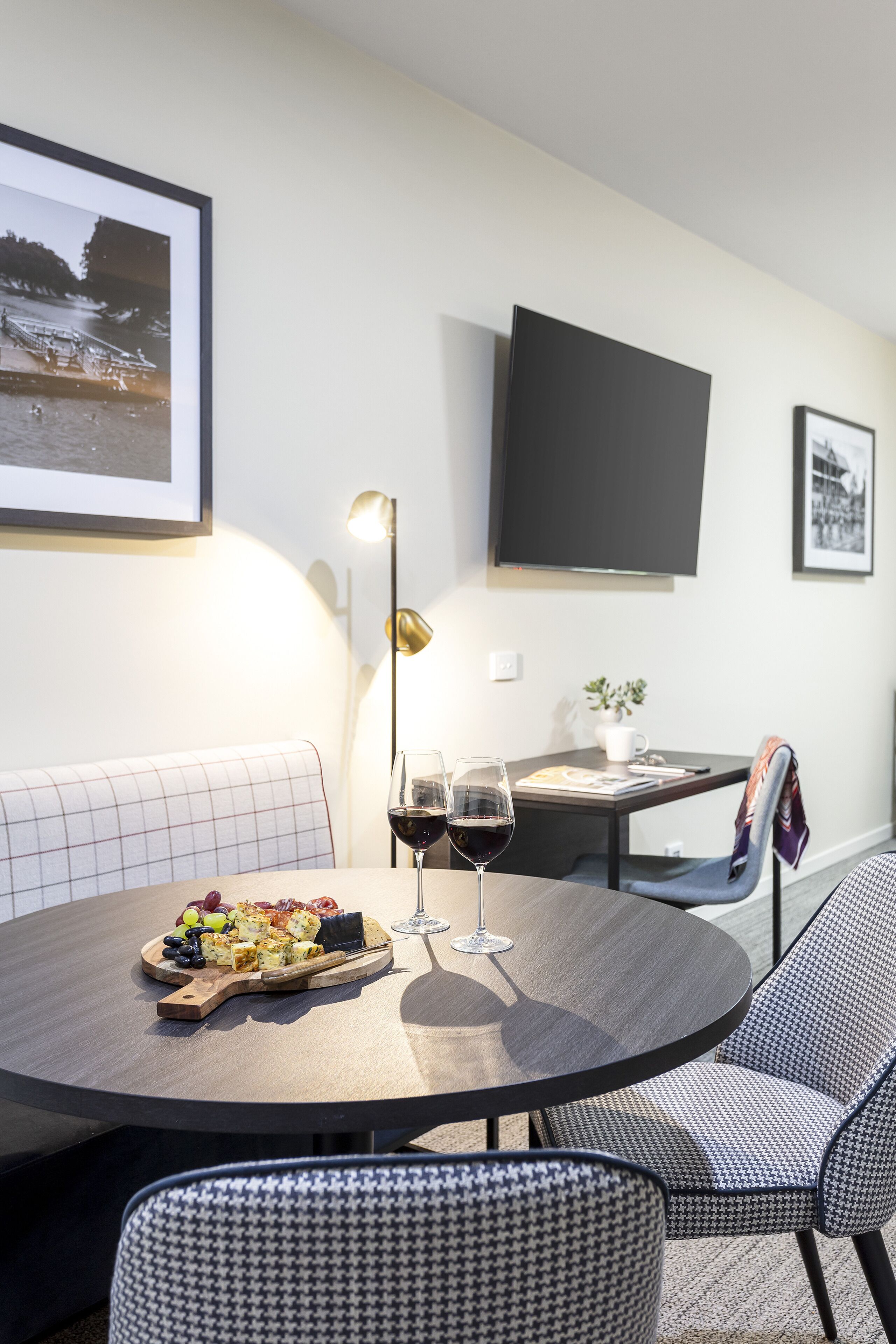 A round dining table with a fruit platter, wine glasses, and a candle. There are two upholstered chairs, a bench, and a wall-mounted TV.