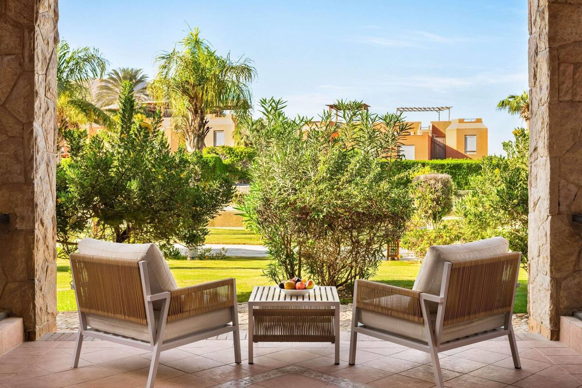 Eine Terrasse mit zwei Korbstühlen und einem kleinen Tisch, mit Blick auf einen gepflegten Garten und ein Gebäude im Hintergrund.