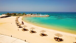 A beach with rows of sun loungers and umbrellas, a clear blue sea, and a distant shoreline with buildings. - Mövenpick Resort El Sokhna (Ataqah)