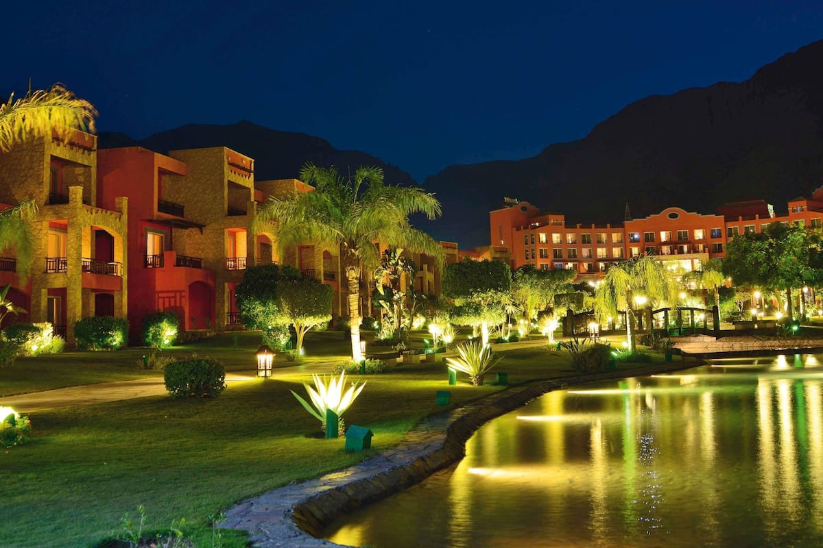 A hotel with multiple buildings, a well-lit garden, and a calm water feature.