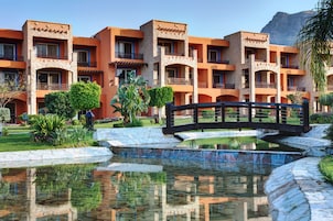 Un hotel con varios balcones, un puente sobre un estanque reflectante y montañas al fondo.