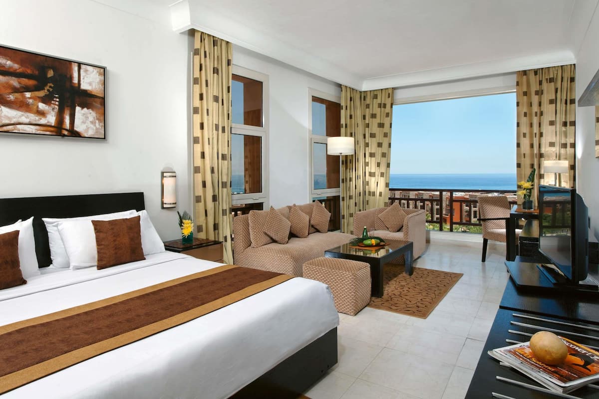 A hotel room with a bed, sofa, armchair, and a balcony with a view of the sea.