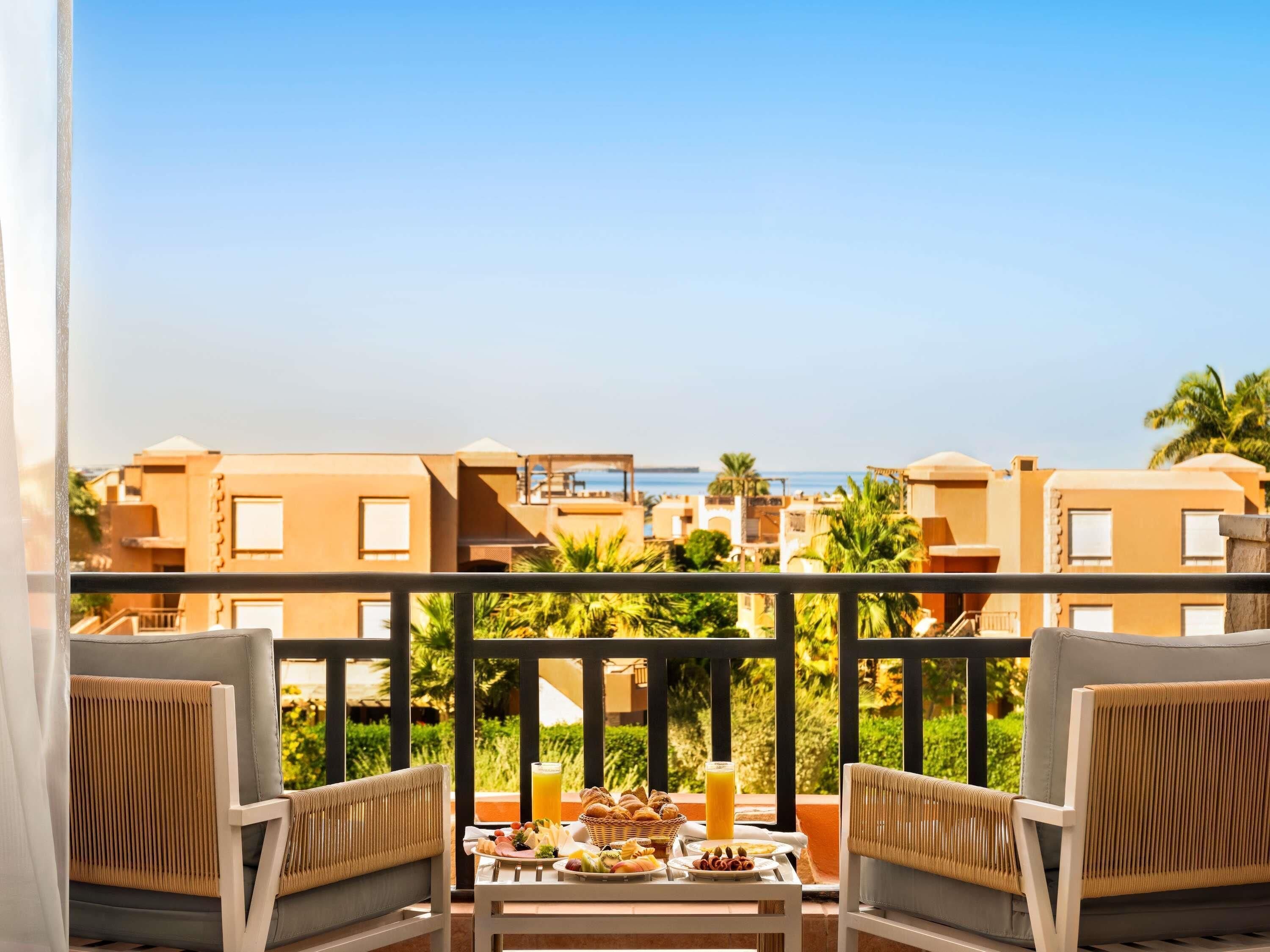 A balcony with two chairs, a table with breakfast, and a view of buildings and the sea.