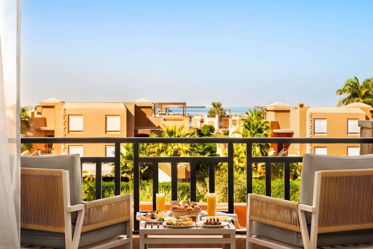 A balcony with two chairs, a table with breakfast, and a view of buildings and the sea.