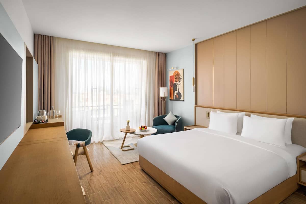 A hotel room with a large bed, a desk with a chair, a small table with a vase, and a window with curtains.