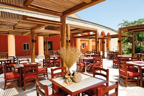 Un área de comedor al aire libre con mesas y sillas de madera, un muro de piedra y un cielo azul despejado.