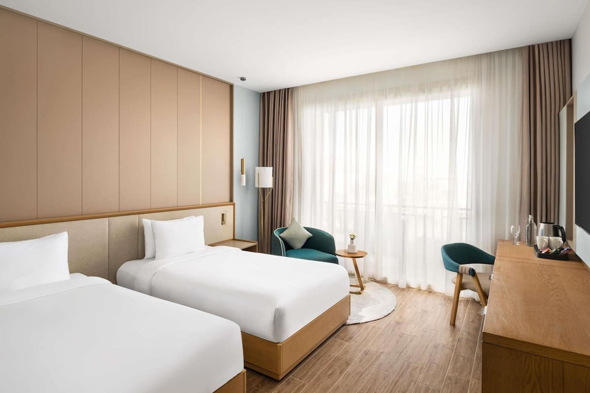 A hotel room with two beds, a desk, a chair, a lamp, and a large window with curtains.