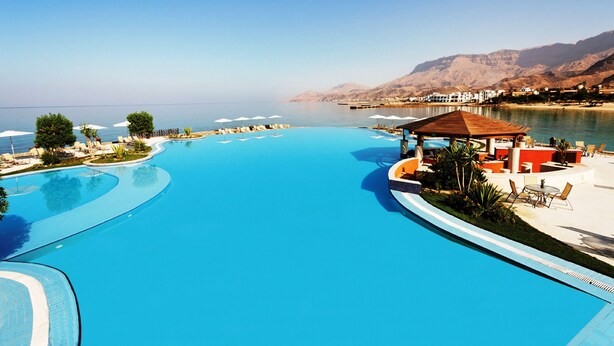 A large, curved swimming pool with lounge chairs and umbrellas, surrounded by mountains and a clear sky. - Mövenpick Resort El Sokhna (Ataqah)