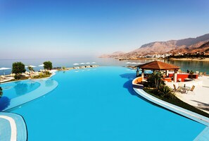 Una gran piscina curva con sillas de descanso y sombrillas, rodeada de montañas y un cielo despejado.